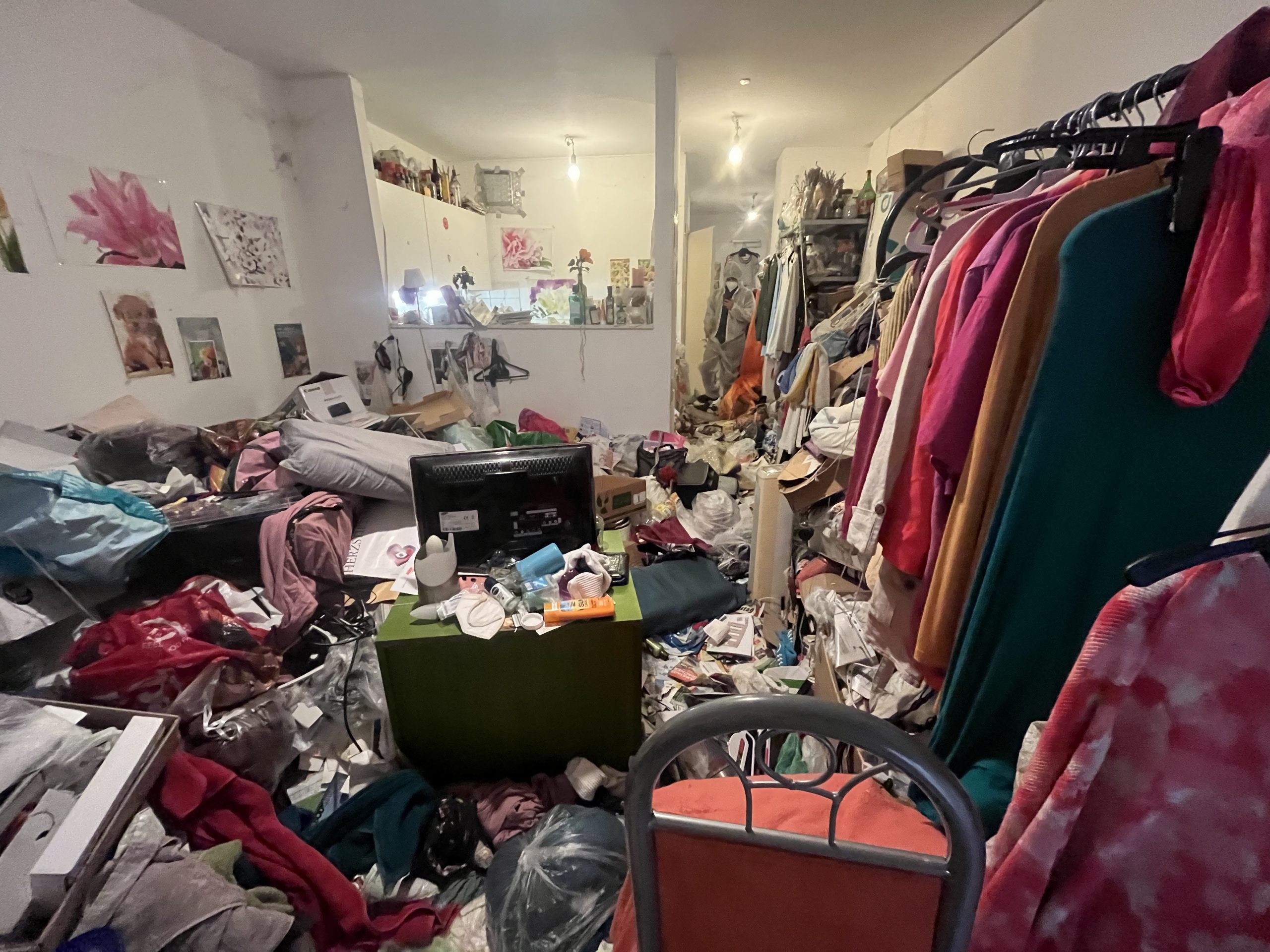 Unordentliches Zimmer, bedeckt mit Kleidung, Müll und verstreuten Gegenständen, auch als 'Messi-Haus' bekannt. Im Hintergrund sind einige Bilder an der Wand zu sehen.