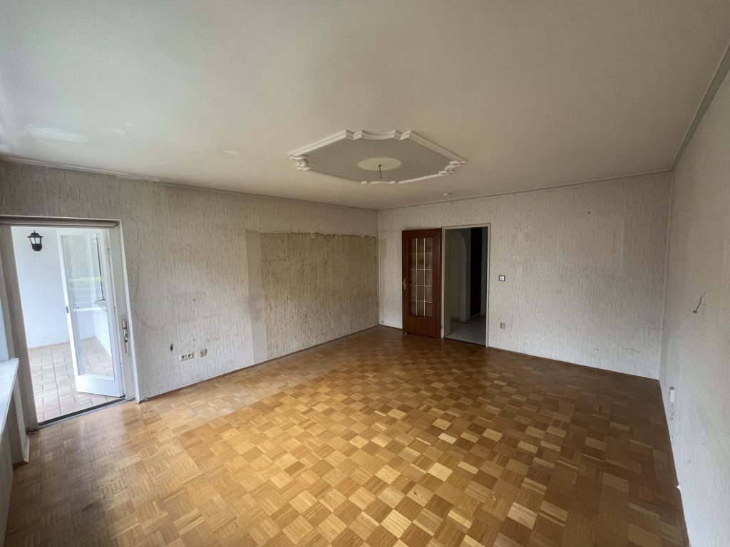 Wohnzimmer vor Renovierung mit alten Tapeten, sichtbaren Wandstellen und Holzboden.