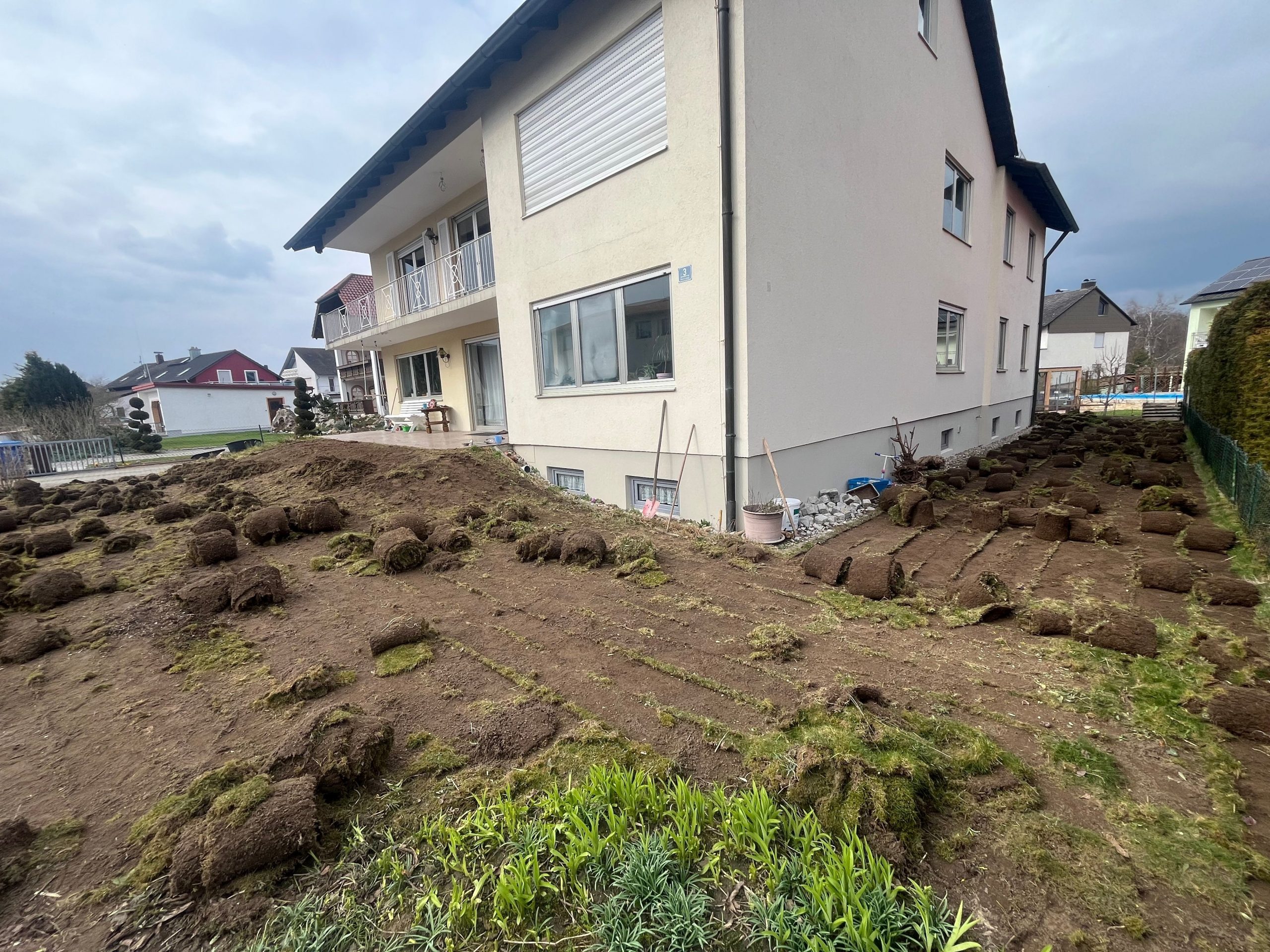 Gartenarbeiten vor einem Mehrfamilienhaus, bei denen alte Grasflächen entfernt und der Boden für neuen Rasen vorbereitet wird.