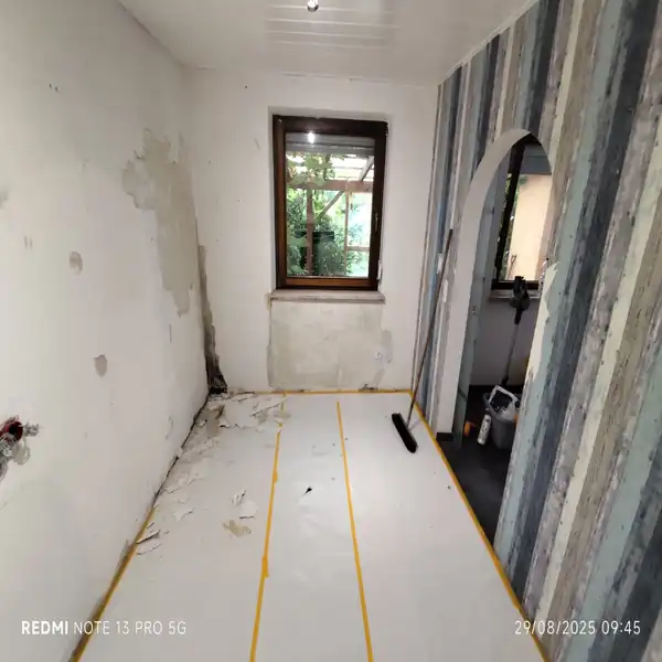 Raum in Renovierung mit entfernten Tapeten, beschädigten Wandflächen und abgedecktem Boden, bereit für weitere Renovierungsarbeiten.